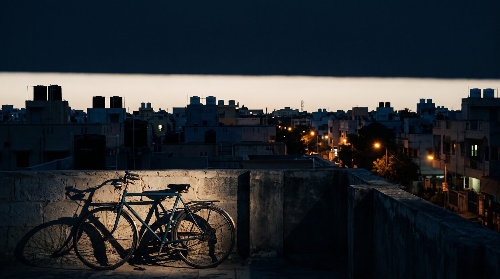 A Chennai rooftop at dusk, with a bicycle against a parapet and amber street lights across the neighbourhood below.